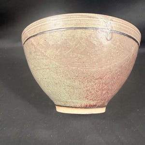 May include: A handmade ceramic bowl with a wide, rounded shape. The bowl features a muted color palette of green, brown, and red hues. A dark band encircles the rim, adding a contrasting detail to the textured surface.
