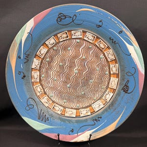 May include: A decorative ceramic plate with a blue rim and a brown and white patterned center. The plate features a wavy design in the center, surrounded by a border of small squares. The rim has colorful accents and black line art.