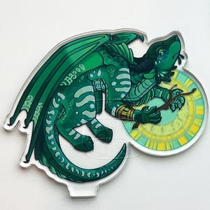 May include: A green dragon-like creature with large wings and a long tail, holding a stick over a circular, yellow-and-green clock face. The artwork is on a clear acrylic base, with intricate details and a fantasy theme.
