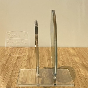 May include: Transparent acrylic display stand with a rectangular base and two vertical supports. One support is a thin, rectangular shape, and the other is a curved, circular shape. The stand is designed to hold a small item. The text "Henix Sketching Bird Studios" is visible.