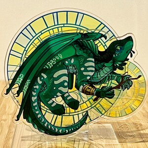 May include: A green dragon illustration with white markings and large wings. The dragon is holding a stick, set against a yellow and white clock-like background. The artwork is on a clear acrylic stand.