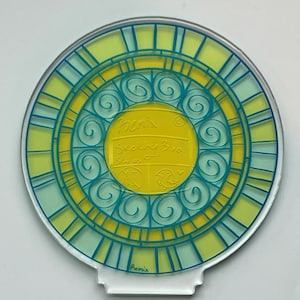 May include: A clear, circular acrylic coaster with a stained-glass design. The coaster features a yellow centre with teal and yellow concentric circles and teal lines. The text "Sketchy Bino Studios" is visible in the centre.