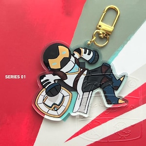 May include: A clear acrylic keyring featuring a cartoon character in a futuristic outfit with a gold-coloured clasp. The character is primarily white, black, and blue, with gold accents. The text "SERIES 01" is visible.
