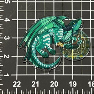 May include: A clear acrylic charm depicting a teal dragon with green wings and details. The dragon is holding a stick in front of a circular design with gold and green details. The charm is on a black grid background.