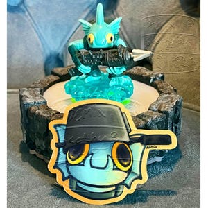 Op de afbeelding: Een turquoise waterwezen-figuurtje dat een donkergrijs wapen vasthoudt, staand op een ronde basis met steentextuur. Vooraan ligt een platte sticker met een gestileerde illustratie van hetzelfde wezen met een zwarte bucket hat met tekst.