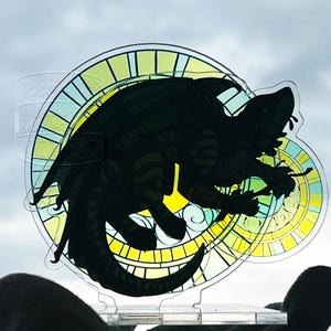 May include: A clear acrylic dragon silhouette with a stained-glass style background in yellow, green, and blue. The dragon is black and detailed, with wings and a long tail. The background has a circular design.