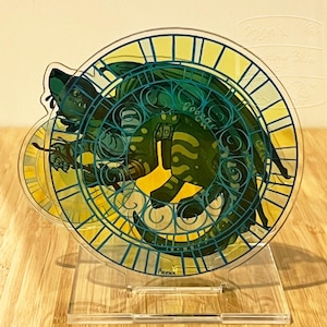 May include: A clear acrylic stand featuring a detailed illustration of a dragon within a circular stained-glass design. The dragon is primarily green and yellow, with blue accents. The stand is on a clear base, and the background is a light wood.