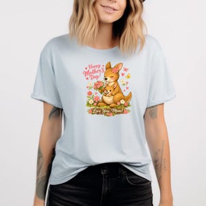 May include: Light blue t-shirt featuring a cartoon kangaroo with a joey, surrounded by flowers and butterflies. The text reads "Happy Mother's Day! Love You, Mom!" in pink and green. The shirt has short sleeves and a crew neck.