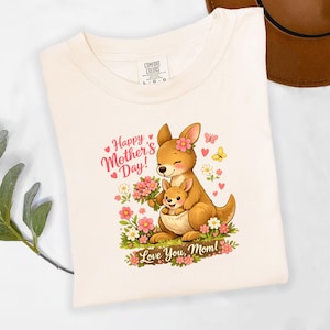 May include: Cream-colored t-shirt featuring a cartoon kangaroo and joey with flowers, hearts, and butterflies. The text reads "Happy Mother's Day!" and "Love You, Mom!". The shirt has a Comfort Colors tag.