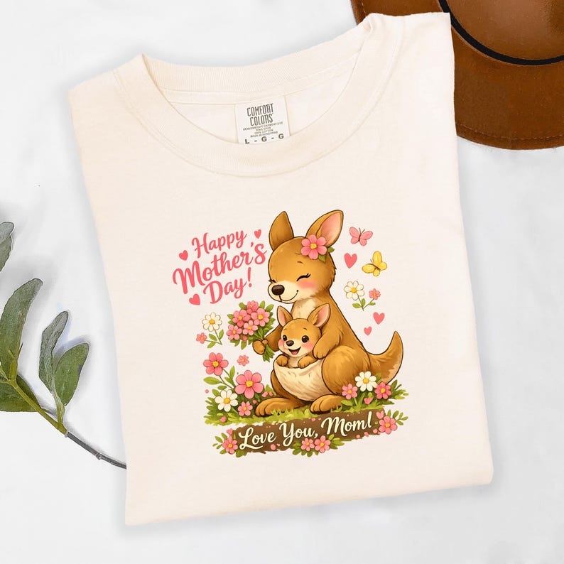 May include: Cream-colored t-shirt featuring a cartoon kangaroo and joey with flowers, hearts, and butterflies. The text reads "Happy Mother's Day!" and "Love You, Mom!". The shirt has a Comfort Colors tag.