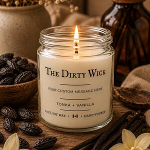 Peut inclure: Une bougie allumée dans un pot en verre transparent avec une étiquette indiquant The Dirty Wick, Your Custom Message Here, Tonka + Vanilla, 100% Soy Wax et Hand-Poured. La bougie possède une mèche allumée et contient de la cire blanche.