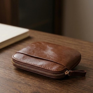 Puede incluir: Una cartera pequeña y cuadrada de color marrón con un acabado liso y pulido y una textura granulada visible. La cartera cuenta con una cremallera metálica de tono dorado en tres de sus lados, con una lengüeta de cuero marrón a juego.