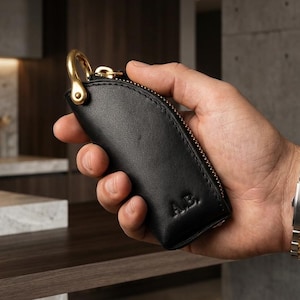May include: Black leather key holder with a gold-coloured zip and clasp. The key holder has the initials "A.B." embossed on the front. The key holder is held in a hand.