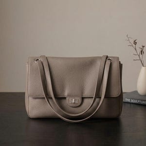 Puede incluir: Un bolso de hombro de cuero color topo con textura granulada, que presenta una solapa rectangular, dos correas largas para el hombro y un cierre giratorio rectangular de tono plateado centrado en el panel frontal.