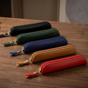 May include: Five slim, cylindrical pencil cases arranged in a diagonal row. Each case features a gold-toned zip with a matching leather pull tab. The colours from back to front are black, dark green, navy blue, tan, and bright red.