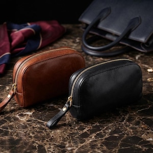 Puede incluir: Dos neceseres de cuero, uno marrón y otro negro, con cremalleras doradas y correas de muñeca. Un bolso azul marino y una bufanda estampada están en el fondo. Los neceseres están sobre una superficie de mármol.