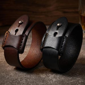 May include: Two leather wrist cuffs, one brown and one black, are shown on a textured surface. Each cuff has a buckle closure with a metal stud. The brown cuff is open, while the black cuff is closed.