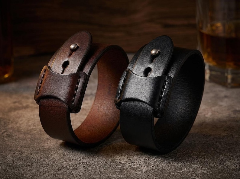 May include: Two leather wrist cuffs, one brown and one black, are shown on a textured surface. Each cuff has a buckle closure with a metal stud. The brown cuff is open, while the black cuff is closed.