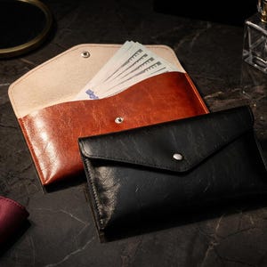 Puede incluir: Dos carteras de cuero, una negra y otra marrón, se exhiben sobre una superficie oscura. La cartera marrón está abierta, revelando una pila de billetes. La cartera negra está cerrada con un botón plateado.