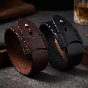 May include: Two leather wrist cuffs, one brown and one black, are shown. Each cuff has a buckle closure with a silver stud. The cuffs are thick and appear to be made of high-quality leather, suitable for a rugged style. The cuffs are approximately 3 cm wide.