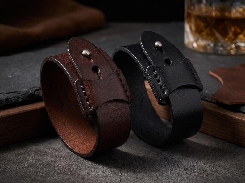 May include: Two leather wrist cuffs, one brown and one black, are shown. Each cuff has a buckle closure with a silver stud. The cuffs are thick and appear to be made of high-quality leather, suitable for a rugged style. The cuffs are approximately 3 cm wide.