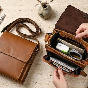 May include: Two tan leather messenger bags with adjustable shoulder straps. One bag is closed, while the other is open, revealing multiple interior compartments containing a smartphone, a small pack of tissues, a circular mirror, and documents.