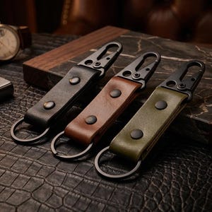 Puede incluir: Tres llaveros de cuero en negro, marrón y verde oliva, cada uno con un mosquetón y un anillo de metal. Los llaveros se exhiben sobre una superficie texturizada, con un reloj y un encendedor en el fondo.