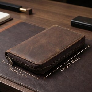 May include: A brown leather zippered case, 18 cm long and 11 cm wide, rests on a dark brown leather desk mat. The case has a distressed finish. Other items on the desk include a black pen, a notebook, and a small black box.