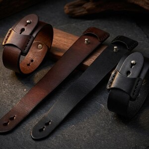 May include: Leather wristbands in shades of brown and black. The wristbands feature a buckle closure and are displayed on a wooden surface. The leather straps have a simple, classic design, suitable for everyday wear.