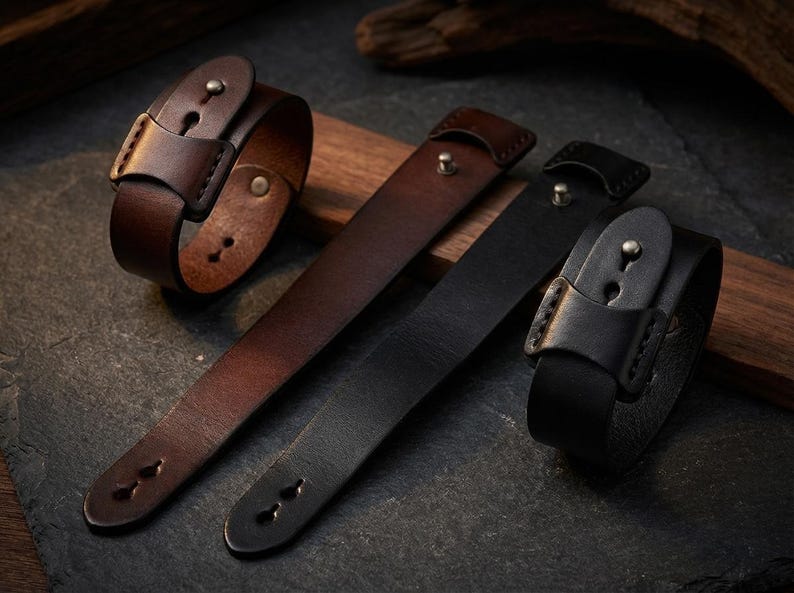 May include: Leather wristbands in shades of brown and black. The wristbands feature a buckle closure and are displayed on a wooden surface. The leather straps have a simple, classic design, suitable for everyday wear.