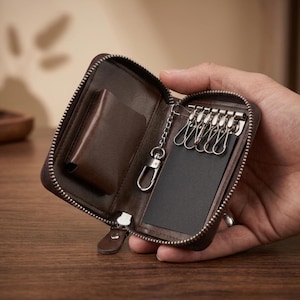 May include: A dark brown leather key case with a zip closure, featuring an interior metal key holder with six hooks and a separate chain with a lobster clasp. The interior includes a small pocket and a black rectangular insert.