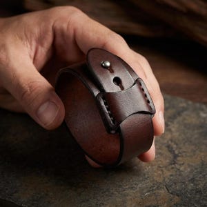 May include: A dark brown leather cuff bracelet with a buckle closure. The bracelet is held in a hand, showcasing the rich colour and texture of the leather. The cuff is approximately 5 cm wide.