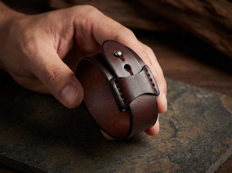May include: A dark brown leather cuff bracelet with a buckle closure. The bracelet is held in a hand, showcasing the rich colour and texture of the leather. The cuff is approximately 5 cm wide.