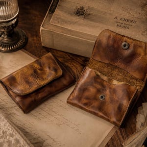 May include: Two brown leather wallets with a distressed finish. One wallet is closed with a flap, while the other is open, revealing a single interior compartment and a metal snap button closure. The leather features visible grain and natural variations.