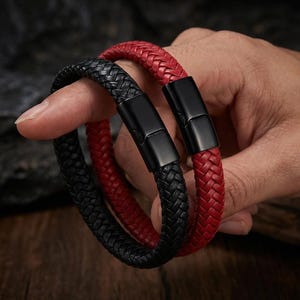 May include: Two braided leather bracelets, one in black and one in red, each featuring a smooth, rectangular black metal magnetic clasp. The bracelets are constructed from multiple strands of leather woven into a consistent, tight pattern.