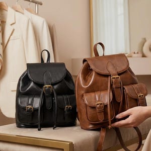 May include: Two leather backpacks, one black and one brown, are displayed. The black backpack has a flap closure and two front pockets with gold-colored buckles. The brown backpack has a similar design. Both have top handles.