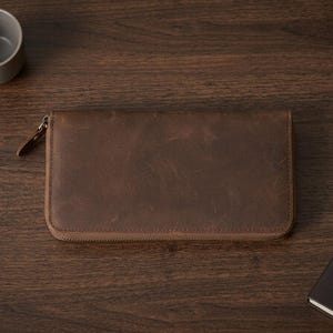 Puede incluir: Una cartera rectangular de cuero marrón con un acabado liso y desgastado, y un cierre de cremallera envolvente. La cartera cuenta con una pequeña lengüeta de cuero unida al tirador metálico de la cremallera, con una textura uniforme.