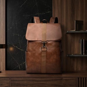 Puede incluir: Una mochila de cuero marrón con cierre de solapa y correas ajustables. La bolsa tiene un aspecto vintage y un cierre de metal. El fondo presenta un panel de madera oscura y una pared de mármol negro.