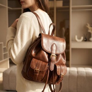 May include: A brown leather backpack with a flap closure and two front pockets. The backpack has adjustable straps and gold-colored buckles. The bag is made of a smooth, shiny leather material.