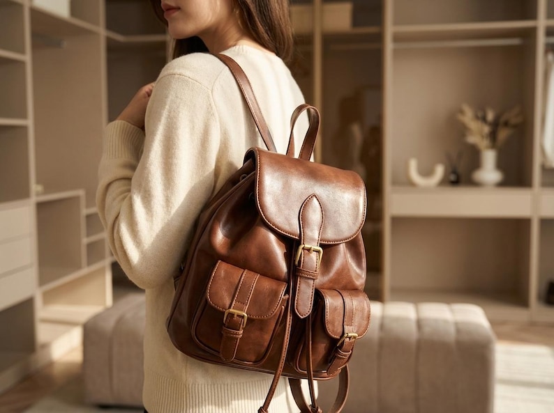 May include: A brown leather backpack with a flap closure and two front pockets. The backpack has adjustable straps and gold-colored buckles. The bag is made of a smooth, shiny leather material.