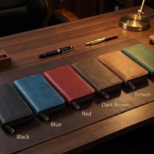 May include: A collection of leather zip-around wallets in various colors, including black, blue, red, dark brown, brown, and green. The wallets are displayed on a wooden desk with pens and a desk blotter.