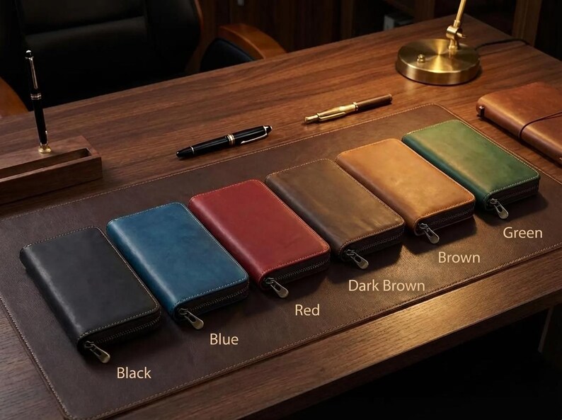 May include: A collection of leather zip-around wallets in various colors, including black, blue, red, dark brown, brown, and green. The wallets are displayed on a wooden desk with pens and a desk blotter.
