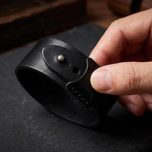 May include: A wide black leather cuff bracelet featuring a smooth surface and a single metallic stud closure. The strap is secured with a small, rounded silver-toned metal button that fits through a pre-cut hole in the leather band.
