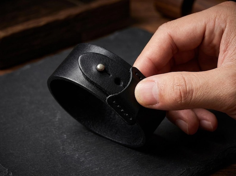May include: A wide black leather cuff bracelet featuring a smooth surface and a single metallic stud closure. The strap is secured with a small, rounded silver-toned metal button that fits through a pre-cut hole in the leather band.