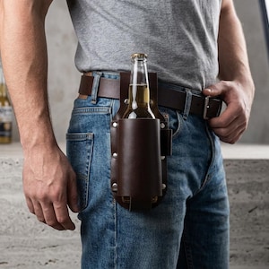 May include: A brown leather beer holster attached to a brown belt, holding a beer bottle. The person is wearing blue jeans and a gray t-shirt. The holster is secured with rivets.