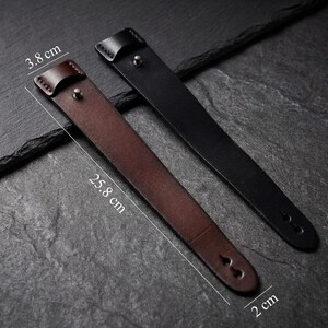 May include: Two leather watch straps, one brown and one black, are shown on a dark surface. The brown strap is 25.8 cm long and 3.8 cm wide, while the black strap is 2 cm wide. Both have a buckle and two holes.