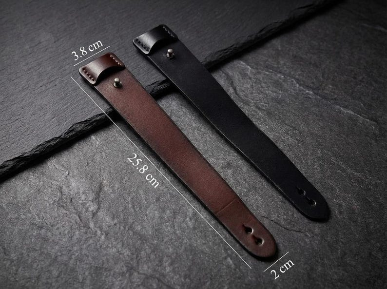 May include: Two leather watch straps, one brown and one black, are shown on a dark surface. The brown strap is 25.8 cm long and 3.8 cm wide, while the black strap is 2 cm wide. Both have a buckle and two holes.