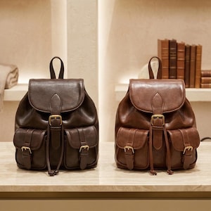 Puede incluir: Dos mochilas de cuero marrón en diferentes tonos. Cada mochila tiene un cierre de solapa, múltiples bolsillos y hebillas doradas. Las mochilas se exhiben sobre una superficie de color claro.