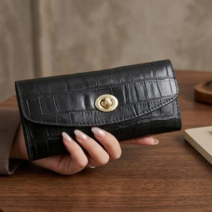 Puede incluir: Una cartera rectangular negra con un patrón de textura de piel de cocodrilo. La cartera presenta una solapa frontal curva asegurada por un cierre giratorio de metal dorado pulido centrado en el panel frontal, ofreciendo un diseño seguro.