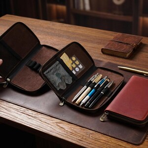 May include: Open brown leather pen case with a zipper, containing several pens. A matching red leather case and a small notebook are also visible. The items are on a brown leather desk mat, with a marble paperweight and a gold pen.
